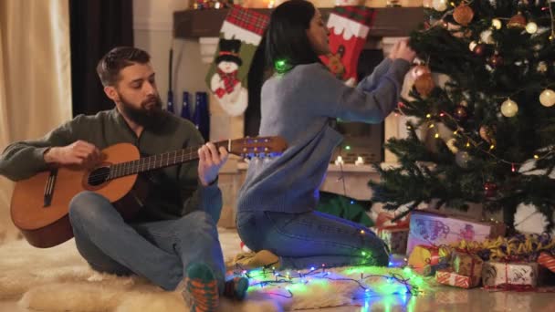 男と女の新年の木に座って 女の子のおもちゃをぶら下げ 男のギターを演奏 クリスマスパーティービデオ — ストック動画