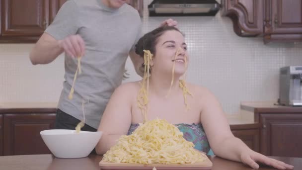 一个胖胖的女人坐在厨房里 男人把面条挂在他胖女朋友的耳朵上 肥胖女人和英俊男人的虚假关系 — 图库视频影像