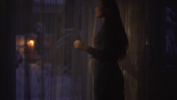 長い金の髪をしたかなり妊娠中の女性が窓に来て 外を見ます 腹を触って笑顔で部屋は暗く ランタンは屋外にあり ビデオ — ストック動画