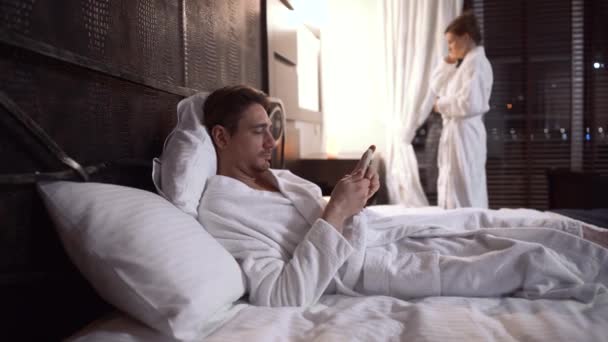 大人の男が快適なホテルで携帯電話にテキストメッセージをベッドに横たわっている 女性の鏡のベッドに行く準備の近くに立っている 幸せなカップルの残りの部分一緒に — ストック動画