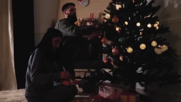 家で新年の木を飾る男と女 女性は小さなギフトボックスに弓を修正します ハッピーカップル準備クリスマスパーティーで暗い部屋 ビデオ — ストック動画