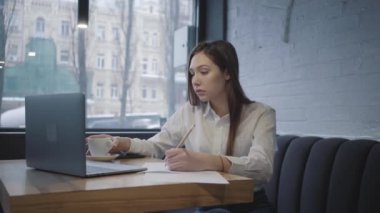 Kafedeki masada dizüstü bilgisayarını kullanan konsantre bir kadın. Düşünceli kız pencerenin yanındaki kanepede oturuyor kahve içiyor ve bilgisayar ekranını kontrol ederek bilgi yazıyor.