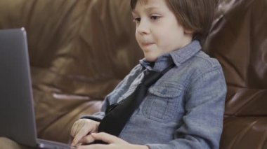 Küçük heyecanlı çocuk kahverengi deri koltukta dizüstü bilgisayarda çalışıyor.