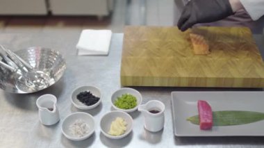 Beyaz restoran üniformalı şefin elleri küçük somon balıklarını kesiyor. Mutfak gereçleri, baharat, kaşıklar, tunbalık parçaları, soslar ve diğer malzemeler..