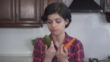 Büyük salatalıkları ısıran ve çiğneyen güzel bir kızın portresi ve mutfakta taze havuç, video. 