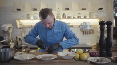 Restoran üniformalı sarışın aşçı ve siyah eldivenli adam modern mutfakta duruyor. Etrafta bir sürü mutfak gereçleri var ve domates kirazı kesiyorlar. Harika yemekler yapmak için masadaki birçok malzemeye.