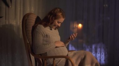 Uzun saçlı hamile bir kadın cep telefonuyla mesaj atıyor ve gülümsüyor. Bayan ekranda göbeğini gösteriyor ve gülümsüyor. 