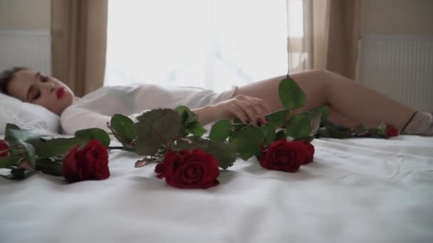 短い透明ナイトウェアでベッドの上に横になって明るいメイクのブロンドの女性のビデオ 女性は花を取り 赤いバラの匂い — ストック動画
