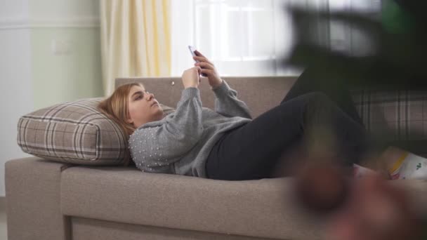 Pufók fiatal nő sms-ezik a mobilján, a kanapén fekszik, közel a nagy ablakhoz. Aranyos plusz méret nő pihentető otthon mobil. A homályos fenyőág az előtérben van. A kamera balra mozog.