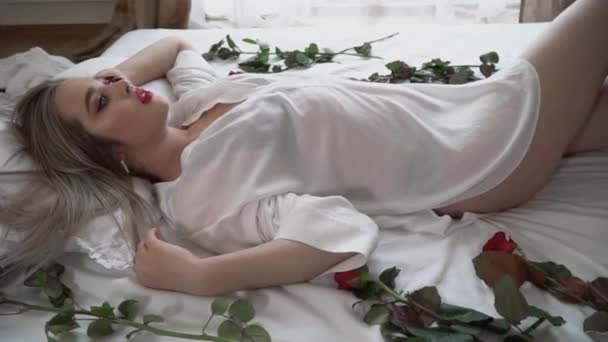透明感のある短いナイトウェアで裏のベッドに横になって明るいメイクの若い女性のビデオ 赤いバラがベッドにいる — ストック動画