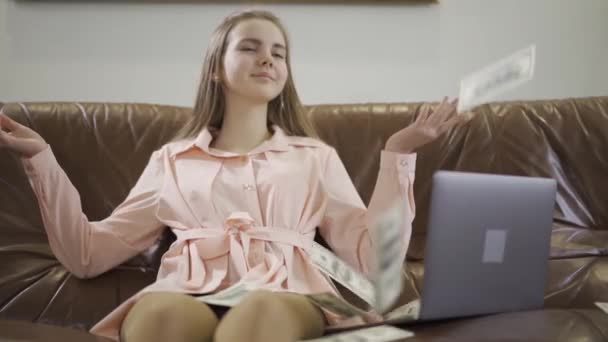 漂亮的少女坐在笔记本电脑前的皮沙发上 美元落在那女孩身上 那孩子面带微笑 很快乐 有钱父母的孩子 慢动作 — 图库视频影像