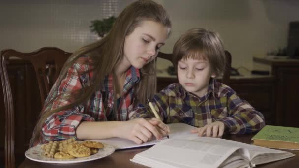 姐姐和弟弟坐在桌旁一起做作业 这个少女帮助一个小男孩做家庭作业 写出正确的答案 爱友好的家庭 — 图库视频影像