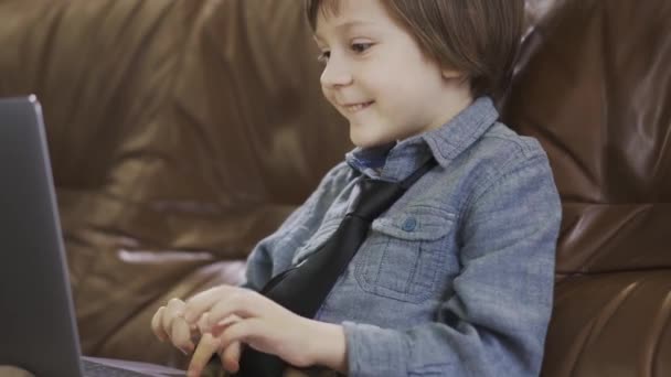 穿着牛仔裤夹克的小男孩坐在皮革沙发上开心地在笔记本电脑上玩耍 现代儿童的闲暇时间 对小玩意上瘾 — 图库视频影像