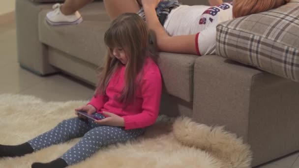 短い赤い髪の若い女性はソファの上に横たわっており 彼女の小さな娘は自宅でふわふわのカーペットの上に座っています 母親と娘は携帯電話にメッセージを女性の世代 — ストック動画
