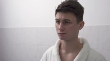 Beyaz bornozlu çekici bir genç adam banyo aynasının önünde duruyor ve saçını düzeltiyor..