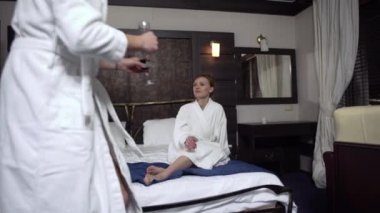 Beyaz bornozlu bir çift geniş bir yatakta oturmuş kırmızı şarap içiyor.