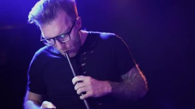 Gözlüklü, nargile tüttüren ve cep telefonuyla gece kulübünde yanıp sönen ışığın arka planına karşı mesaj atan çekici bir genç..