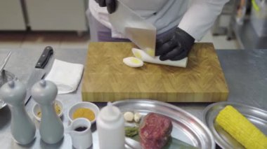 Şefin elleri siyah lateks eldivenlerle kesme tahtasındaki limon çimlerini kesiyor. Mutfak masasında mutfak malzemeleri ve malzemeler var. Mısır, dana eti, sosis, sarımsak, farklı şişeler, baharatlar..