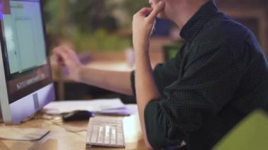 Gözlüklü genç adam çalışma masasında bilgisayar ve video ile oturuyor.  