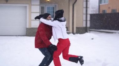 Kışlık elbiseli sakallı bir adam kız arkadaşının etrafında dönüyor, ikisi de gülüyor. Mutlu genç çift kışın arka bahçede eğleniyor.