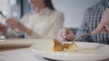 Modern kafede yemek yiyen tanınmayan bir çift. Ön plandaki adam çatal ve bıçakla turta yiyor, arka planda bulanık kadın figürü var. Erkek ve kadın restoranda birlikte vakit geçiriyorlar.