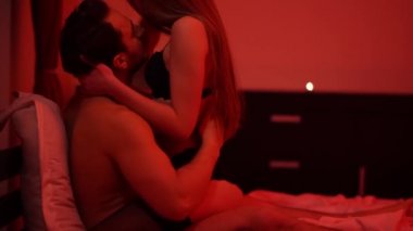 Siyah iç çamaşırlı zayıf bir kadın kırmızı ışıkta yatakta yetişkin bir adamın üzerinde oturuyor. Ateşli çift birbirlerinin vücutlarını okşar, tutkulu ve şefkatle okşar.