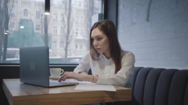 Kafedeki masada laptopunu kullanan düşünceli bir kadın. Pencerenin yanındaki kanepede oturan bir fincan kahve ve bilgisayarla laptopun ekranını kontrol eden bir kız.