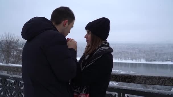 美しい若い女性と男の冬服で川の近くの橋の上に立っている 男は彼らの上に呼吸する女性の手を暖める ビデオ — ストック動画