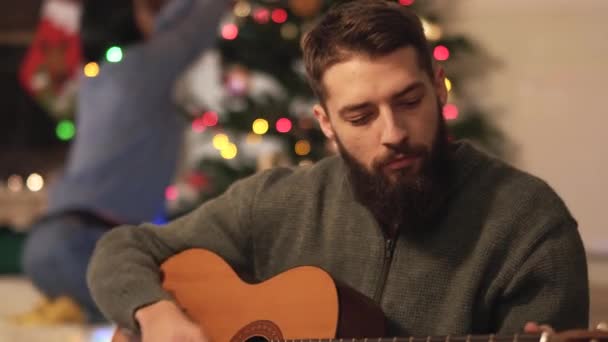 一个留着胡子的男人坐在圣诞树旁的地板上弹奏吉他和唱歌 — 图库视频影像