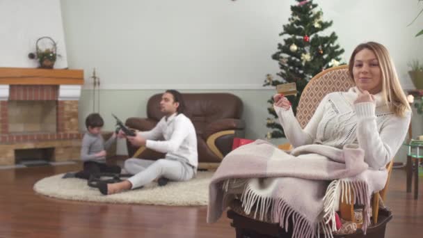 かなりの女性のアームチェアクレジットカードを保持に座っている 背景の床に座っている無人機で遊んでいる父と小さな息子 幸せな家族は家でクリスマスを祝う — ストック動画