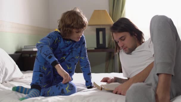 两个兄弟躺在床上 大英俊的哥哥给小可爱的弟弟看了一本书 他们喜欢拍照 — 图库视频影像