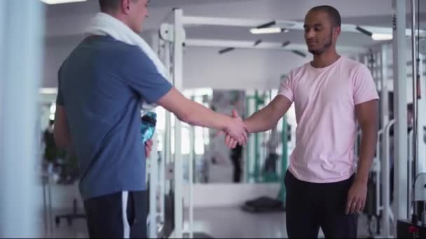 アフリカ系アメリカ人の男が体育館で白人男性と握手をする ジムに２人の若い男がいるコーチ — ストック動画