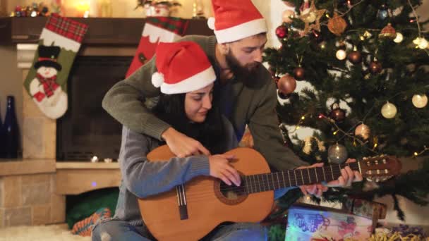クリスマスの前夜にサンタの帽子で幸せなカップルの肖像画 男は女性にギター ビデオを教える — ストック動画