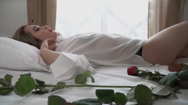 透明感のある短いナイトウェアでベッドに横になって明るいメイクの若い女性のビデオ 赤いバラは女性の体の近くのベッドにあります 若いです女性ルックスDefiantとエロ — ストック動画