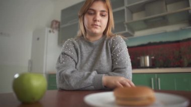 Tombul genç kadın lezzetli bir hamburger ya da sulu yeşil bir elma yemesi gerektiğini düşünüyor. Sağlıklı ve sağlıksız yiyecekler arasında zor bir seçim..
