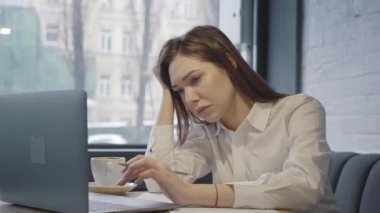 Üzgün genç bir kadın, rahat bir kafe veya restoranın geniş bir penceresinin yanında laptopuyla çalışırken uyuyakalıyor..