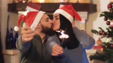 Erkek ve kadın Noel ağacının yanında oturuyor, elinde maytap tutan ve öpüşen kadın, video. 