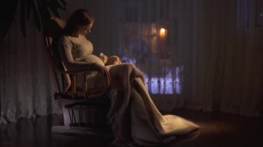 Uzun saçlı güzel hamile kadın karanlık odada sallanan sandalyede oturuyor. Bayan karnını okşa. Kadının bacakları battaniyeyle örtülmüş. Kadın yumuşak oyuncağa sarılıyor, video... 
