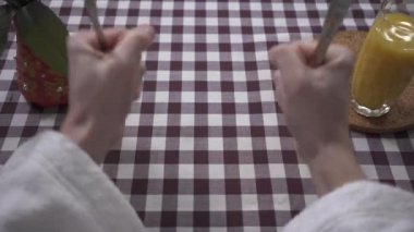 Adamın eli masaya yumruklarıyla vuruyor, yemek istiyor, çatal ve bıçağı yakından tutuyor. Portakal suyu bardağı ve vazo masanın üzerinde kareli masa örtüsüyle birlikte. Beslenme kavramı. İlk ateş eden kişi.