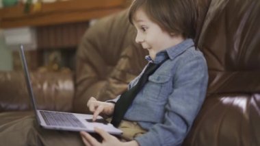 Deri koltukta oturan, dizüstü bilgisayarda oynayan kot ceketli küçük çocuk ekranda şaşırtıcı bir şekilde gülümsüyor. Modern çocuğun boş vakitleri. Aygıtlara bağımlılık