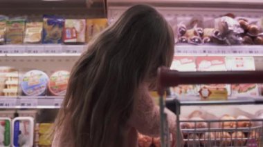 Küçük kız süpermarketteki arabaya sosis koyar. Küçük kız mağazadan ürünleri seçiyor. Marketten alışveriş.