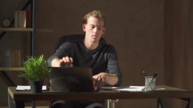 Genç sarışın iş adamı modern ofiste rahat deri koltukta oturur, çalışma yerini düzenler ve evine gider..