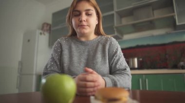 Tombul genç kadın lezzetli bir hamburger ya da sulu yeşil bir elma yemesi gerektiğini düşünüyor. Sağlıklı ve sağlıksız yiyecekler arasında zor bir seçim..