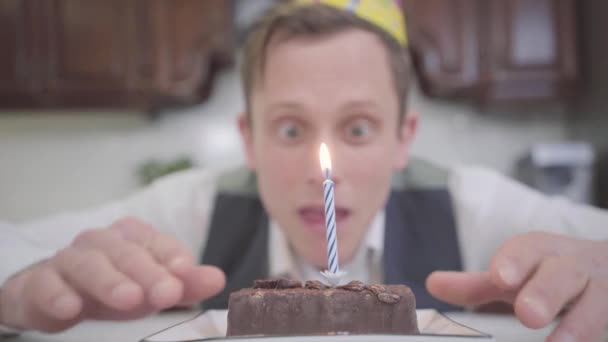 キッチンで小さなチョコレートケーキの前に座って誕生日の帽子で面白い若い男のぼやけた肖像画 オタクは誕生日のお祝いビデオを持っている — ストック動画