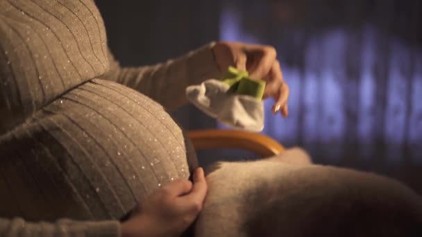未確認の妊娠中の女の子座っていますロッキングチェアで暗い部屋でと遊びます小さな赤ちゃん靴下でそれらを踏みつけます彼女のおなかの上に ビデオ — ストック動画