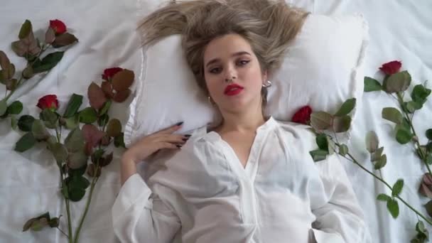 透明パジャマ姿でベッドに横になって明るいメイクの若い女性の動画 赤いバラは体の近くです 若い女性は反抗的でエロティックに見えます レディは遊び心のある気分で彼女の体に触れている — ストック動画