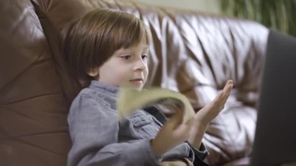 穿着牛仔裤夹克的小男孩坐在手提电脑前的皮沙发上玩钱 孩子手里拿着一捆钱 现代孩子的闲暇时间 富有父母的可爱的快乐儿子 — 图库视频影像