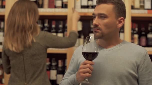 Portrét pohledného muže stojícího se sklenkou červeného vína v obchodě s alkoholem zblízka. Žena v pozadí si vybírá láhev vína. Místo se spoustou alkoholických nápojů.