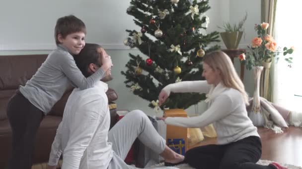 美しい女性は大きな部屋で床に座って彼女の夫にクリスマスプレゼントを与えます 息子は手で父の目を覆う 3人の家族は自宅でクリスマスを祝う — ストック動画