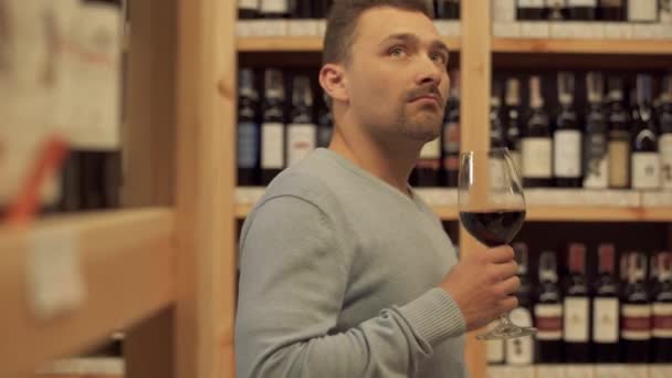 アルコールショップで赤ワインのガラスで立ってハンサムな男の肖像が閉じます 背景の女性はワインボトルを選んでいます 木製の棚にアルコール飲料がたくさんあります — ストック動画